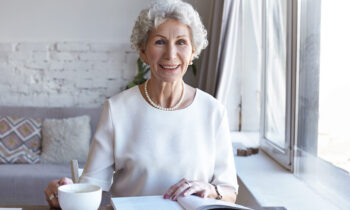 Horizontal shot of cheerful happy retired European gray haired lady relaxing indoors with cup of fresh hot coffee and interesting book. Charming female pensioner reading article in fashion magazine