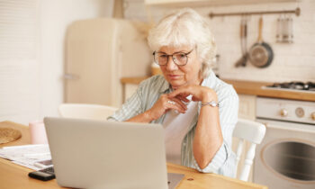Modern technology, old people and retirement. Gray haired female pensioner in glasses filling in loan application form online, sitting in front of laptop, reading information with serious focused look